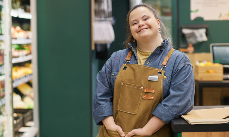 Eine junge weiße Mitarbeiterin mit Downsyndrom im Supermarkt im Kassenbereich lächelt.
