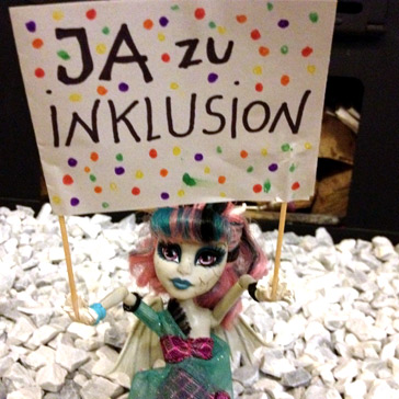 Puppe aus Hartplastik hält ein Plakat in beiden Händen: Ja zu Inklusion.