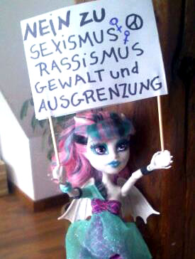 Puppe aus Hartplastik hält ein Plakat in beiden Händen: Nein zu Sexismus, Rassismus, Gewalt und Ausgrenzung
