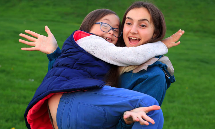 Zwei lachende Mädchen. Ein Mädchen mit Down-Syndrom. Das Mädchen mit Down-Syndrom sitzt auf dem Arm von dem anderen Mädchen.